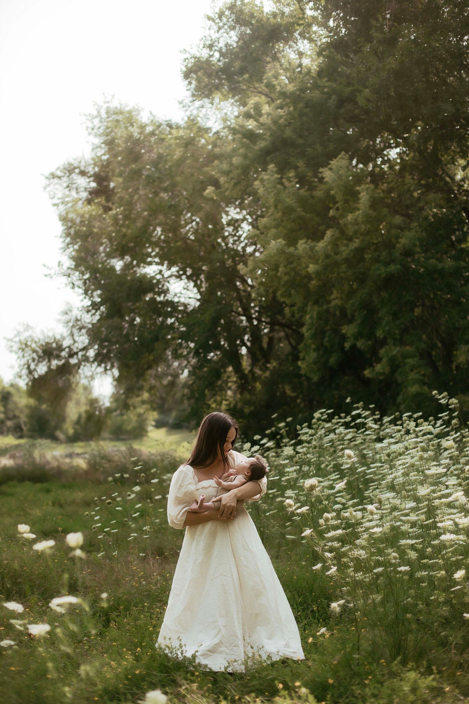 Woman in white dress holding newborn baby in a sunlit meadow with wildflowers and lush green trees.