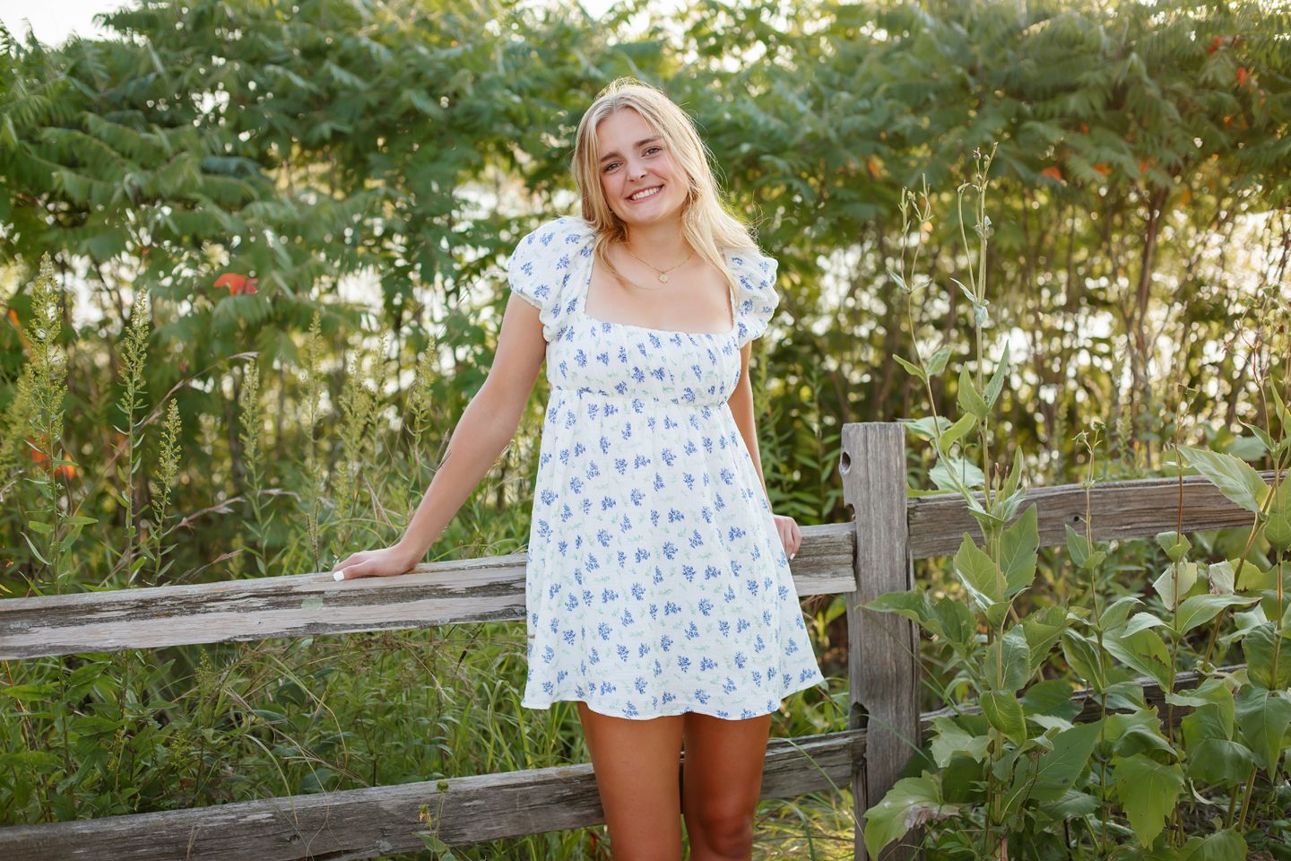 A person in a white floral summer dress leans against a rustic wooden fence surrounded by greenery during golden hour.