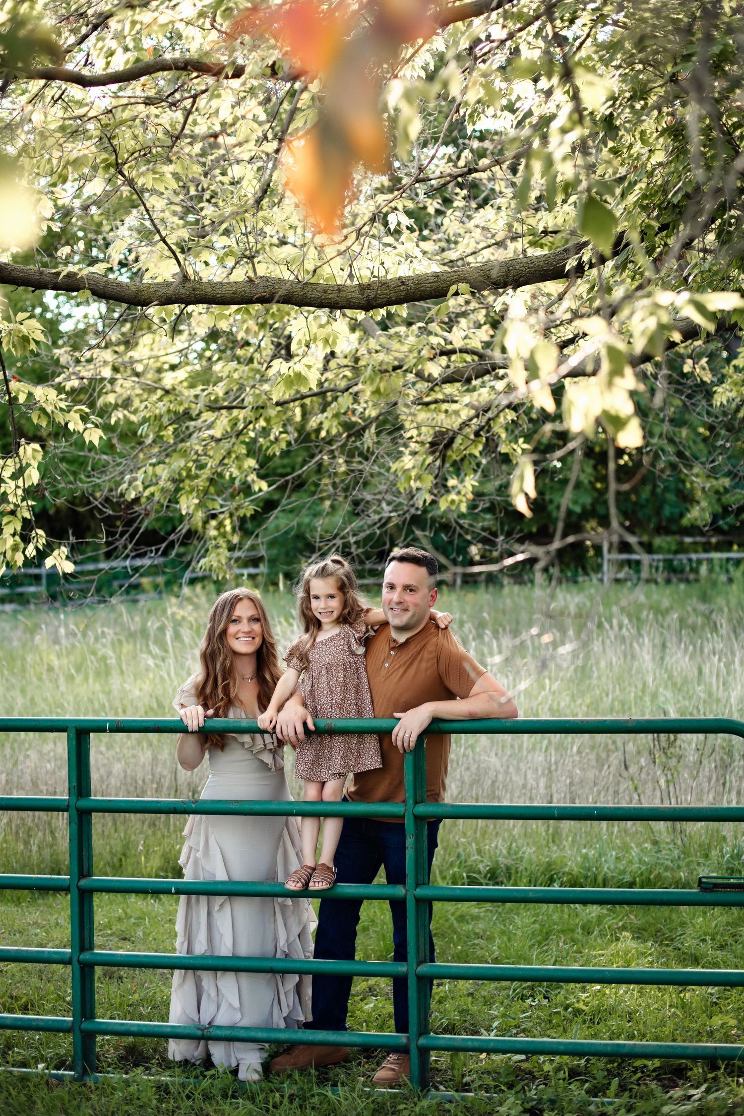 Family standing together at green metal fence in rustic outdoor setting with tall grass and trees in background.
