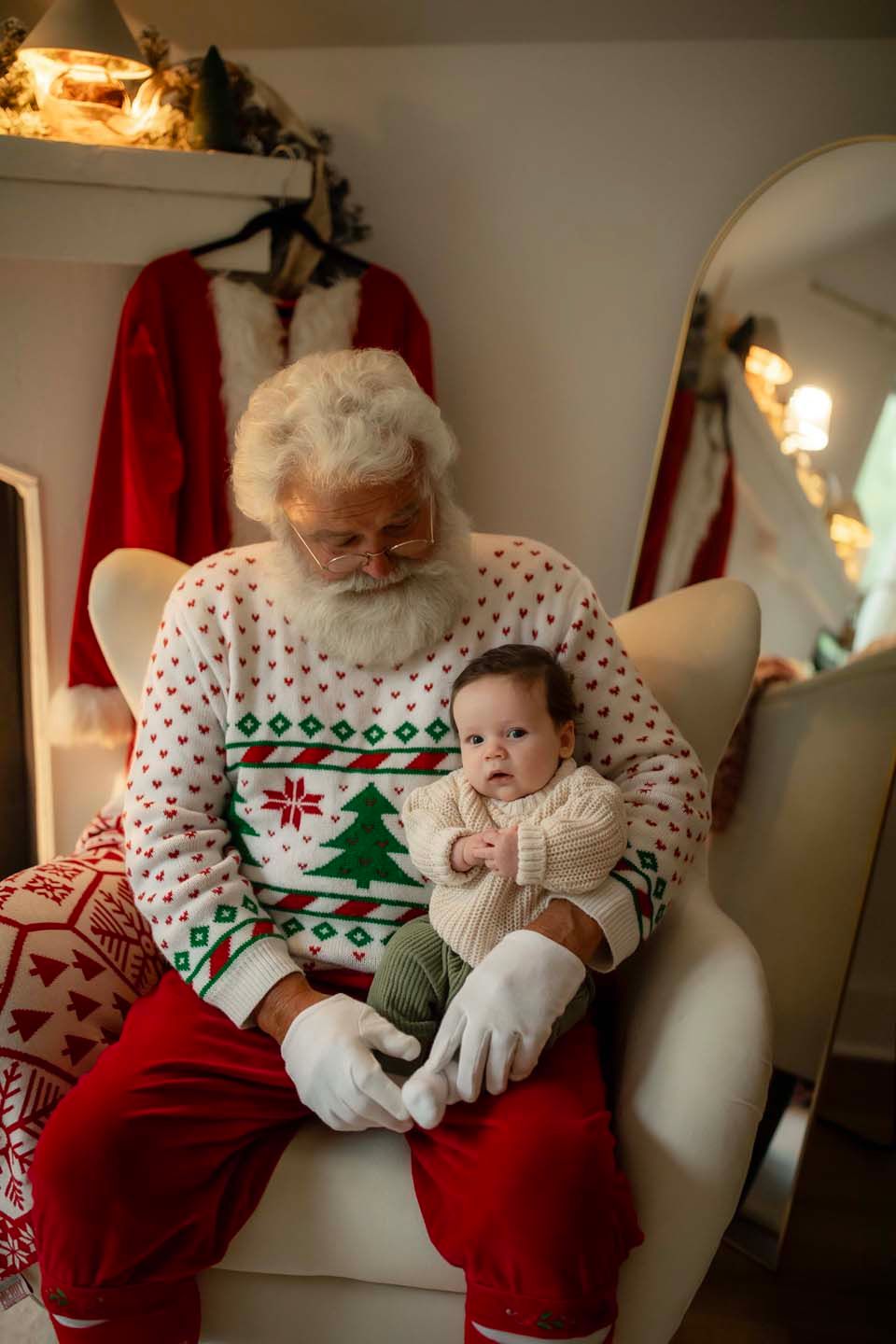 Santa Claus in a Christmas sweater holds a cute baby infant on his lap in a festive holiday setting.