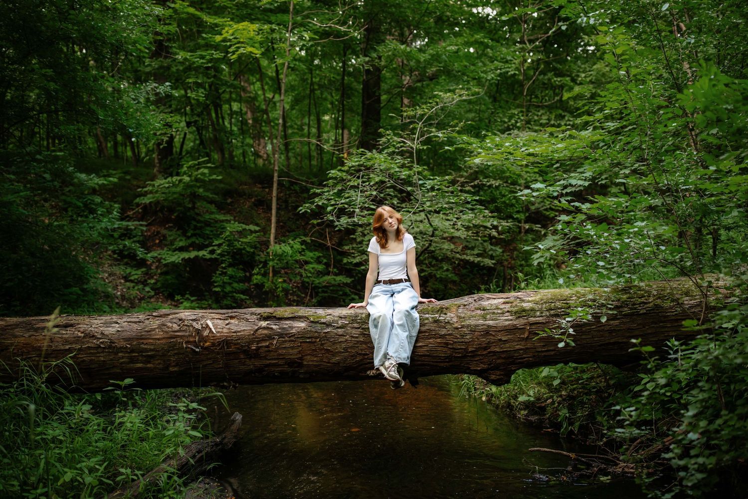 A figure in a white dress sits on a fallen log in a lush green forest setting, creating a dreamy woodland scene.