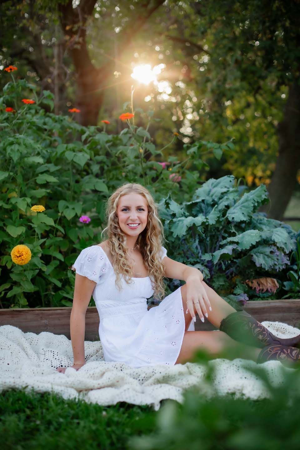 White dress portrait in garden with sunburst through trees and marigold flowers on blanket at sunset.