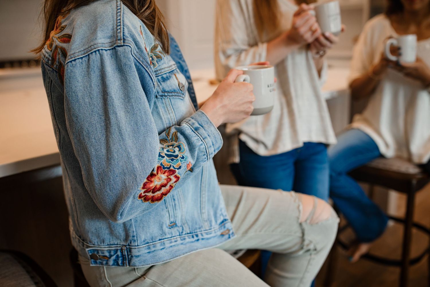 Person wearing embroidered denim jacket and white top in casual indoor setting.