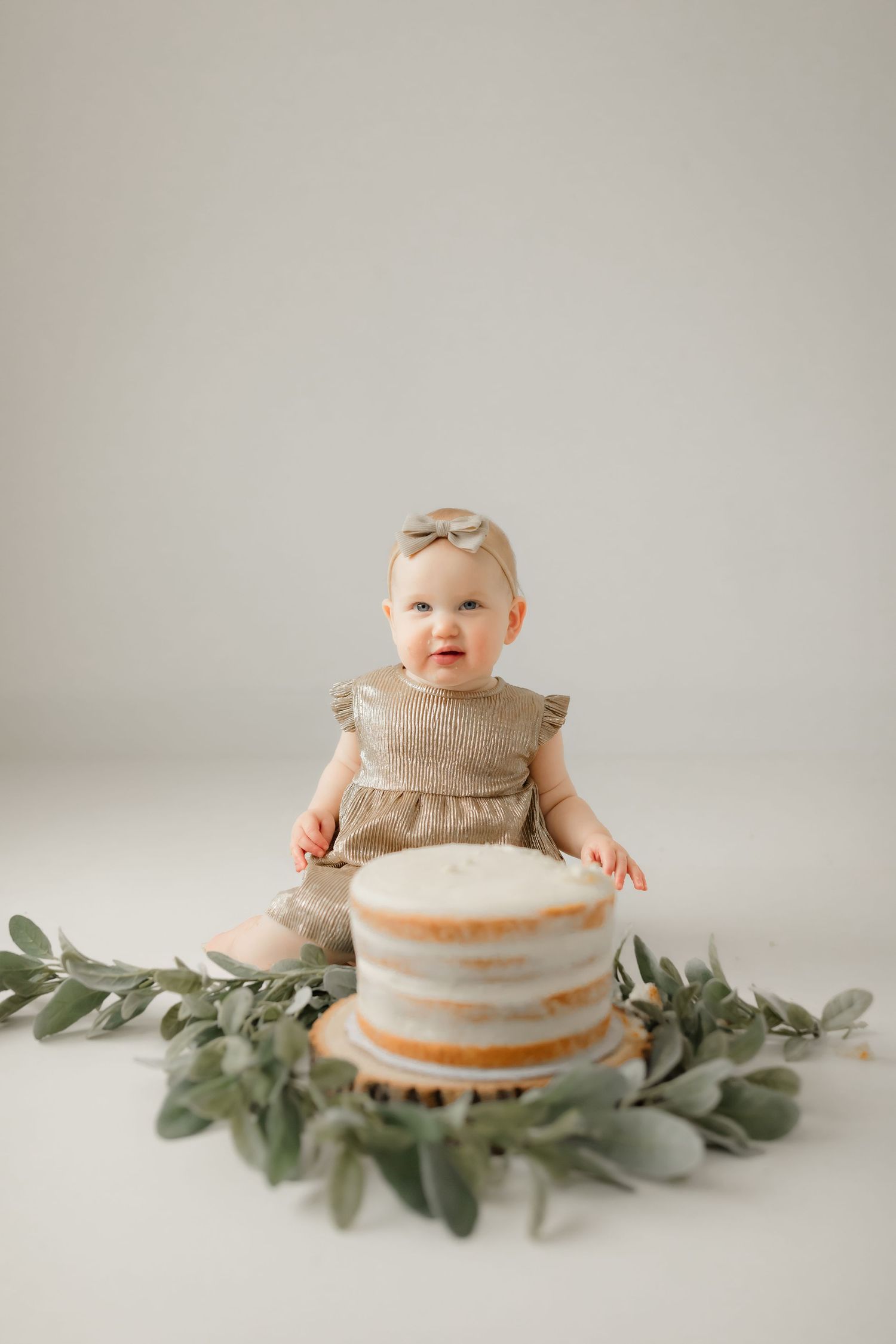 Baby in neutral dress sits by white tiered cake decorated with eucalyptus.