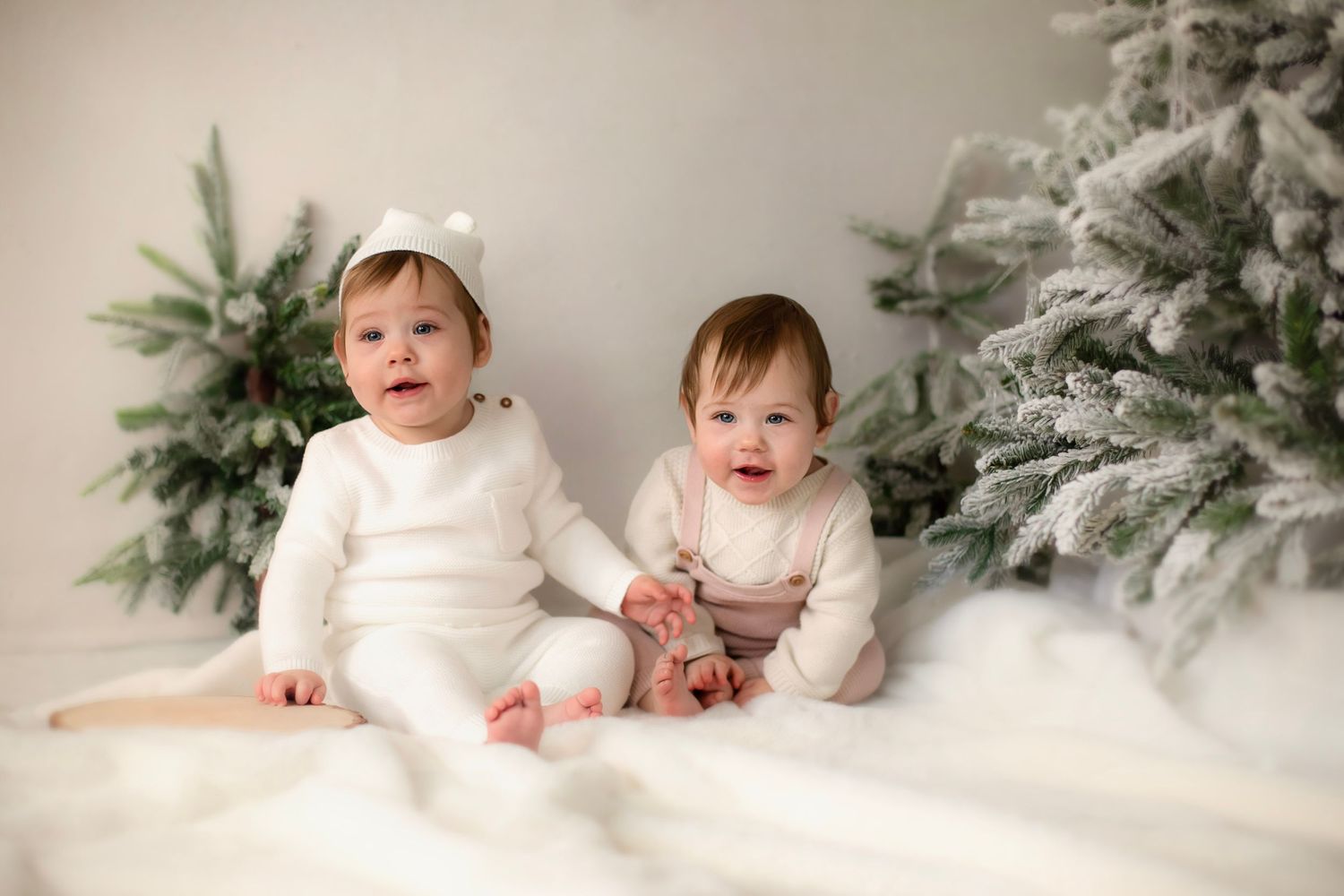 Two babies in white winter outfits sitting on fluffy rug with snowy Christmas trees in background.