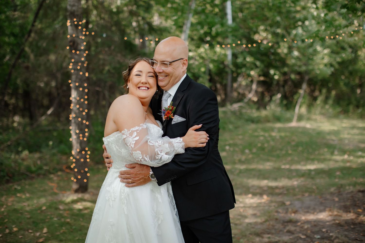 Wedding couple sharing a joyful moment outdoors with string lights and greenery in the background.