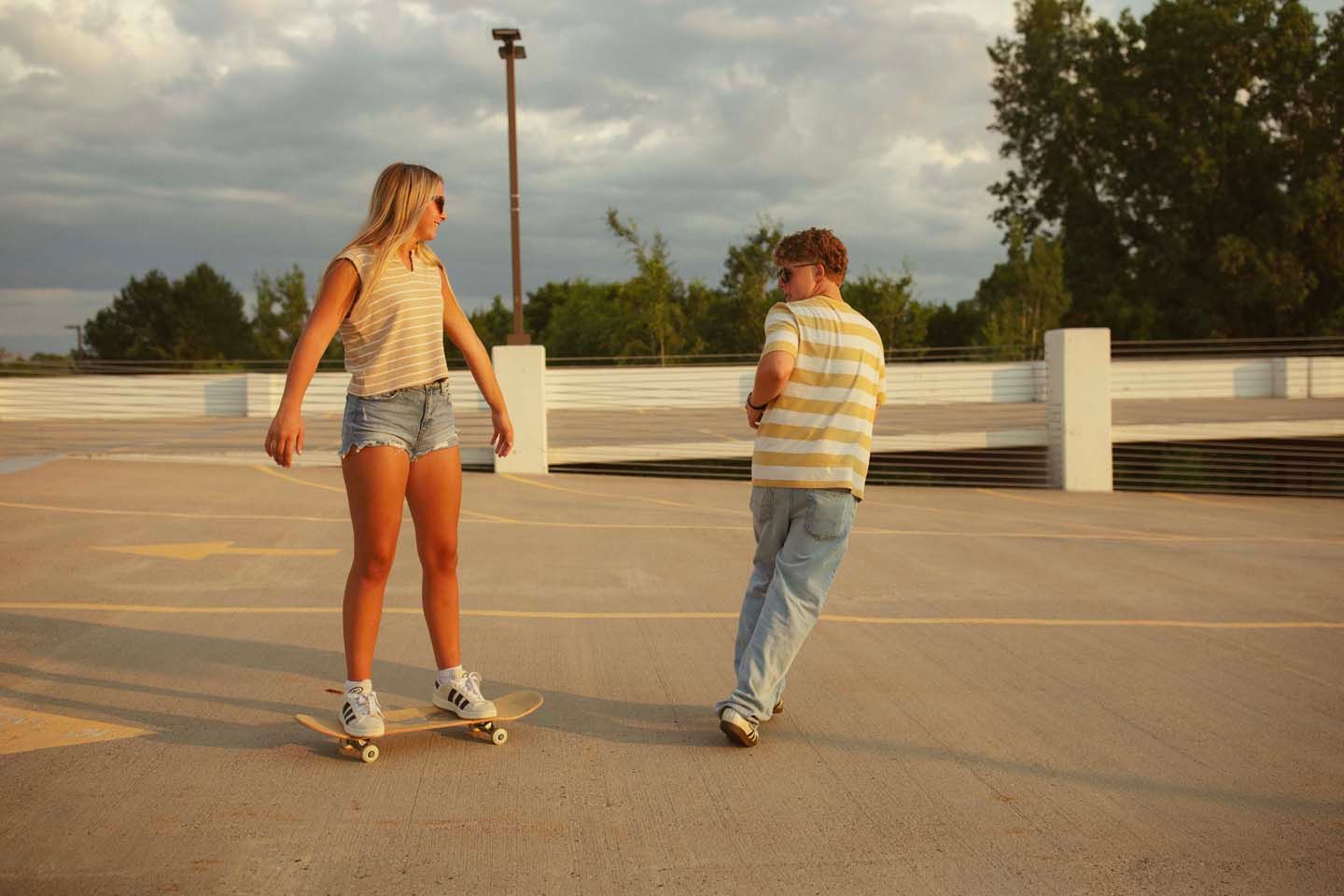 Two young people skateboarding in a sunny parking lot, wearing casual summer outfits, with trees and cloudy sky in background.