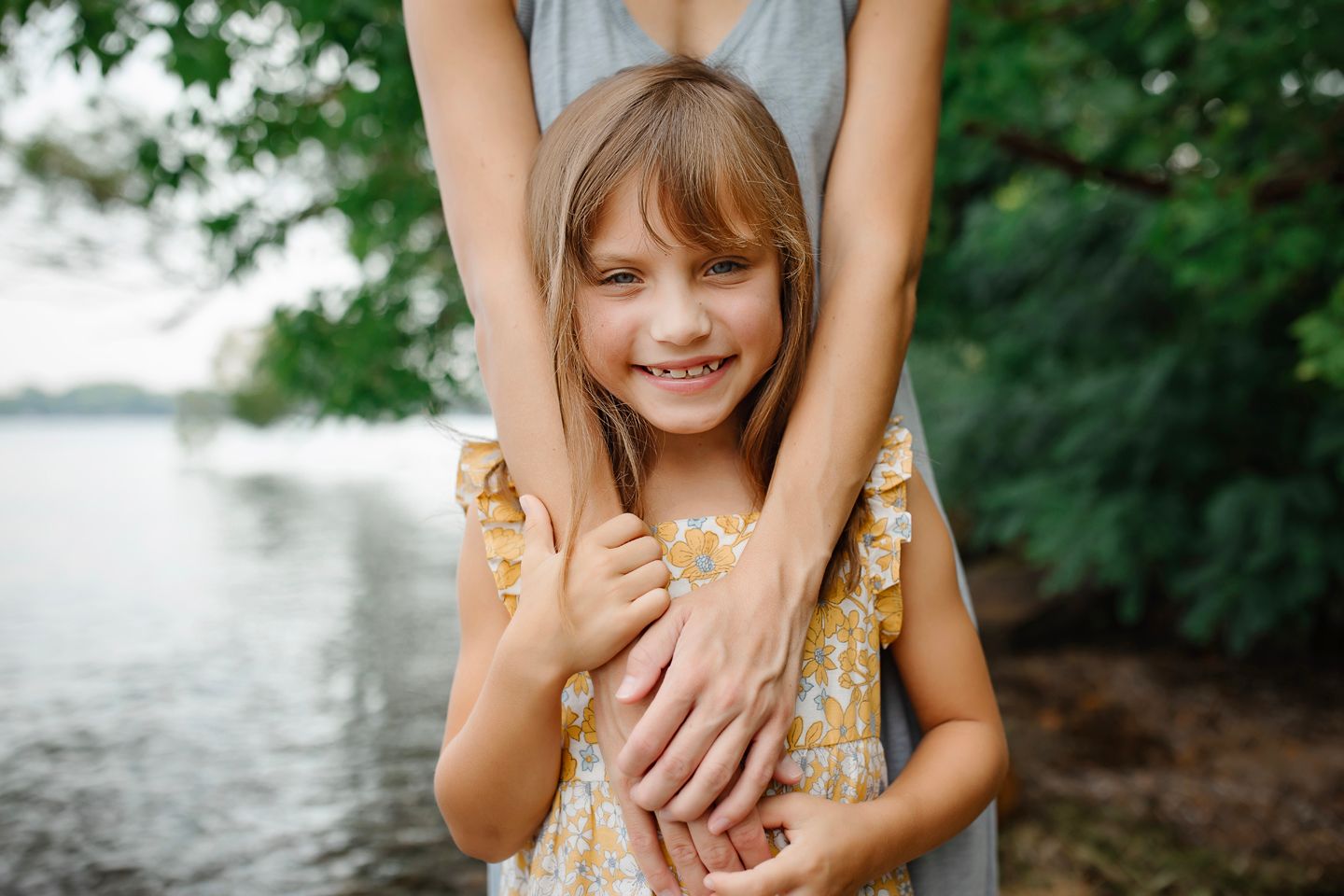A joyful child in a floral dress stands by the lake while adult hands rest on their shoulders.