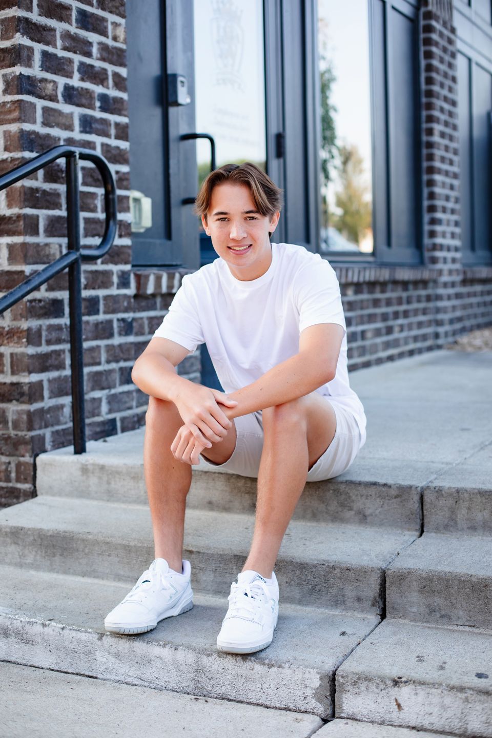 Someone in casual white t-shirt and shorts sits on concrete steps outside a brick building wearing white sneakers.