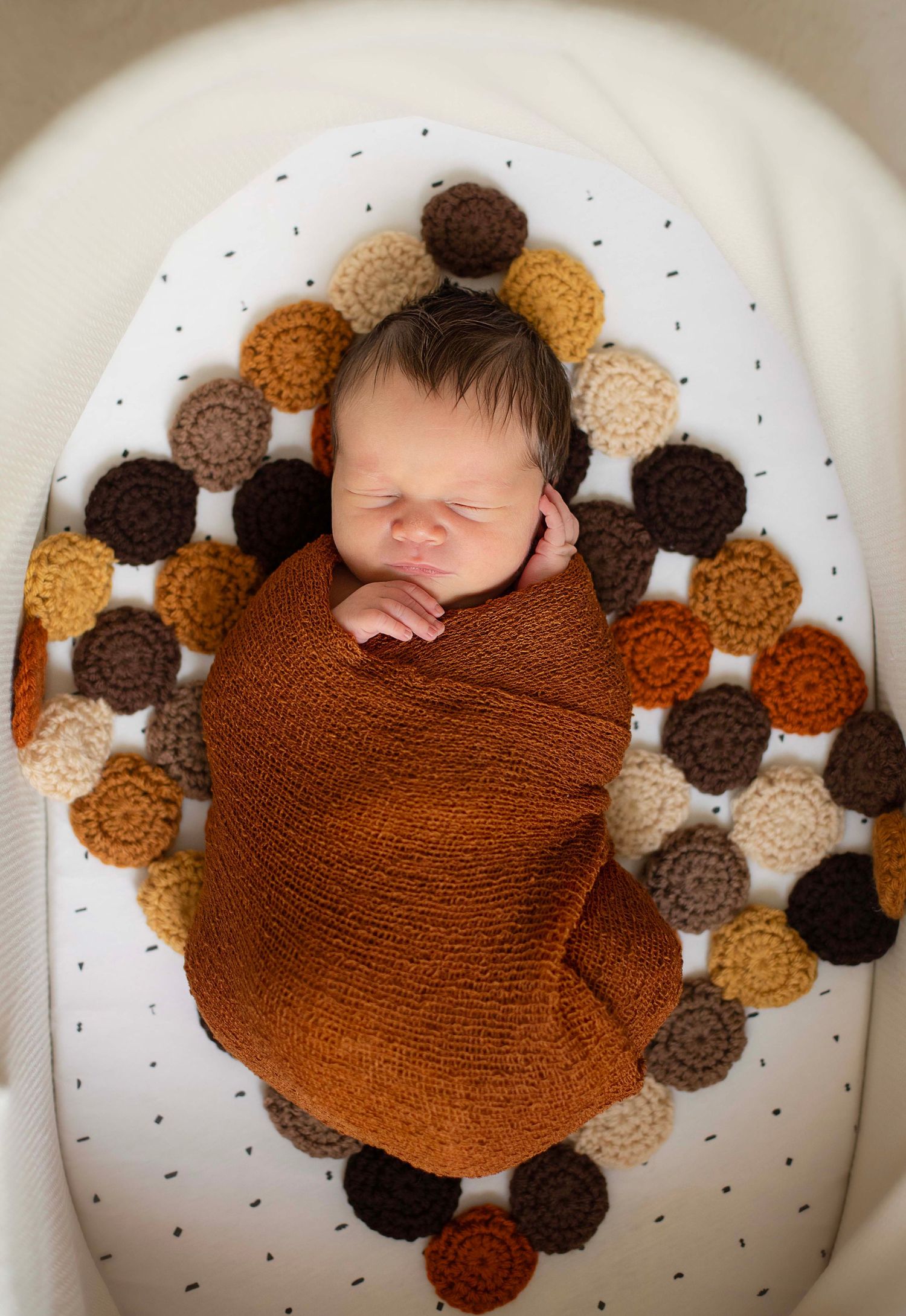 Newborn baby swaddled in rust orange wrap, surrounded by crocheted circles in fall colors, sleeping peacefully in white bassinet.