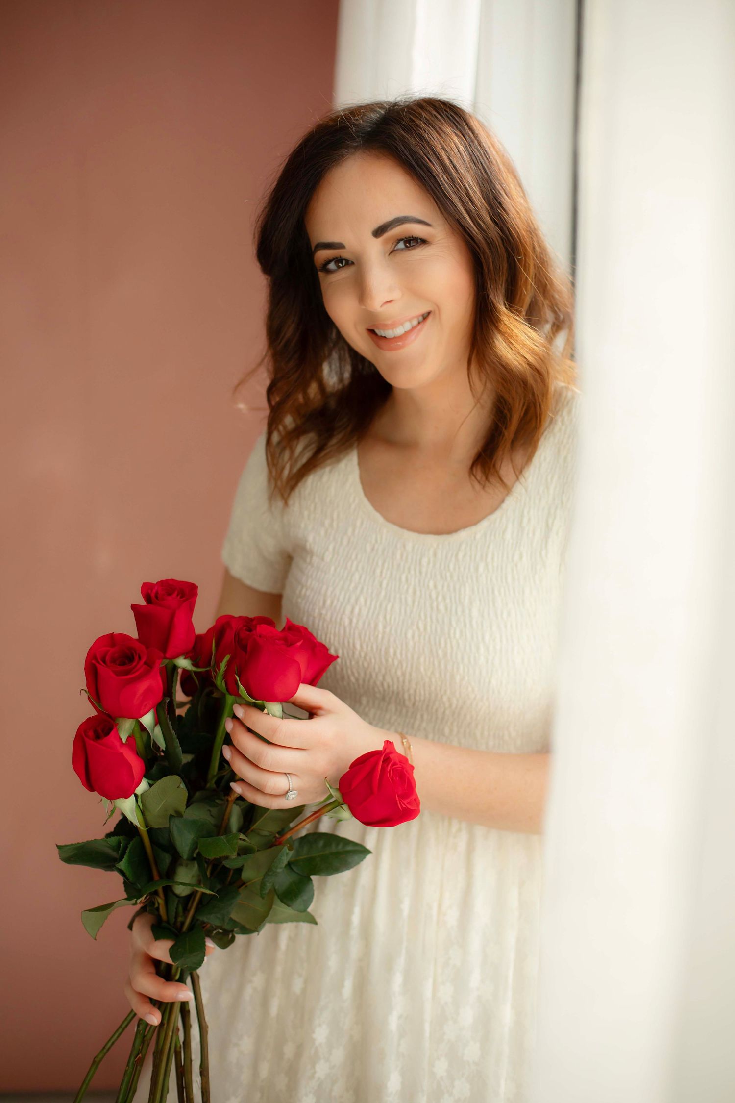 Person in a white dress holding a bouquet of vibrant red roses stands by a window with soft natural lighting.