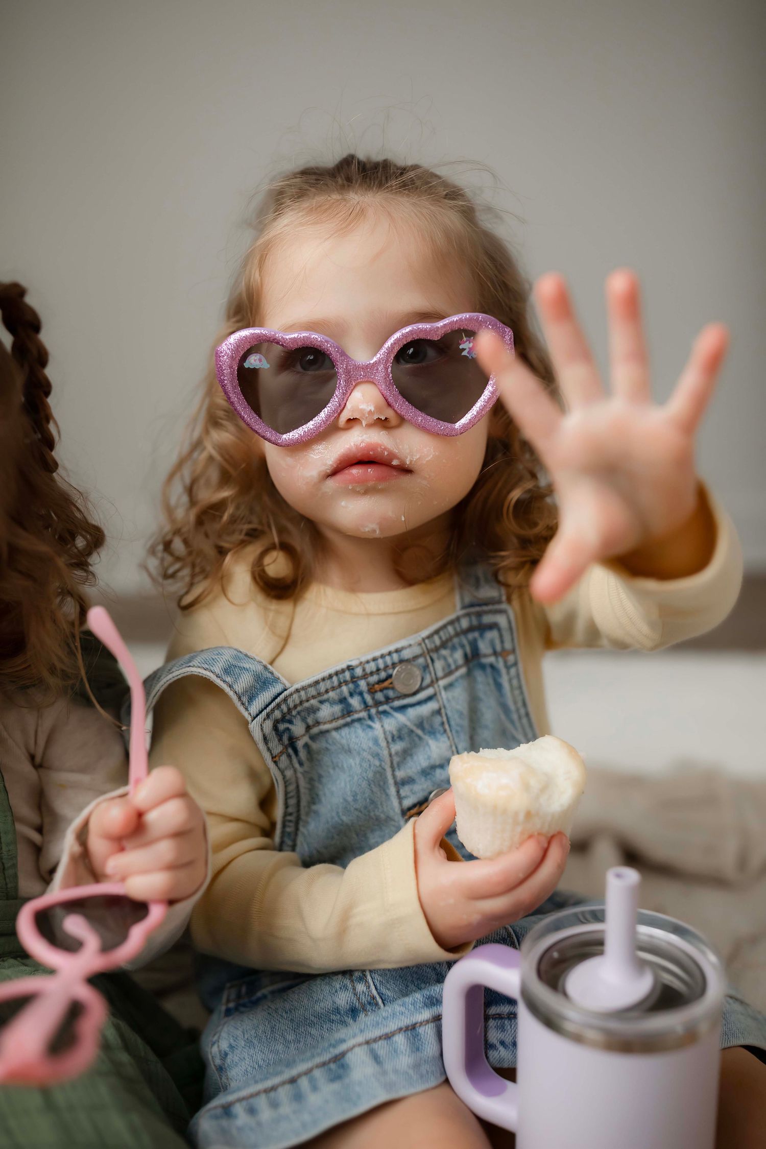 Playful toddler wearing pink sunglasses and overalls reaches toward camera while holding a cup with bendy straw.