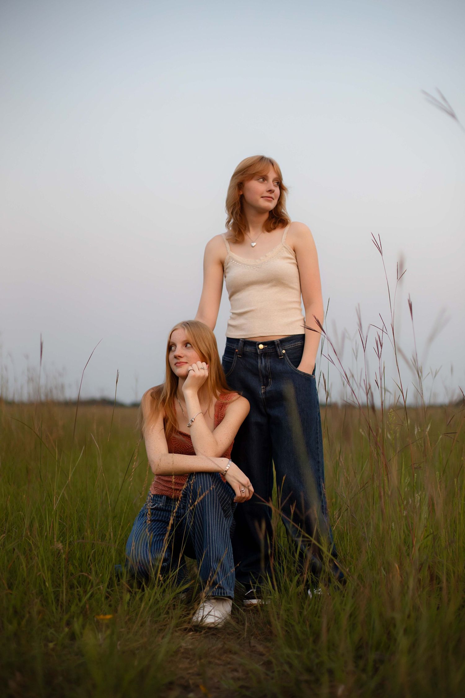 Two young women posing in a grassy field at dusk, one sitting and one standing, both wearing jeans and tank tops.