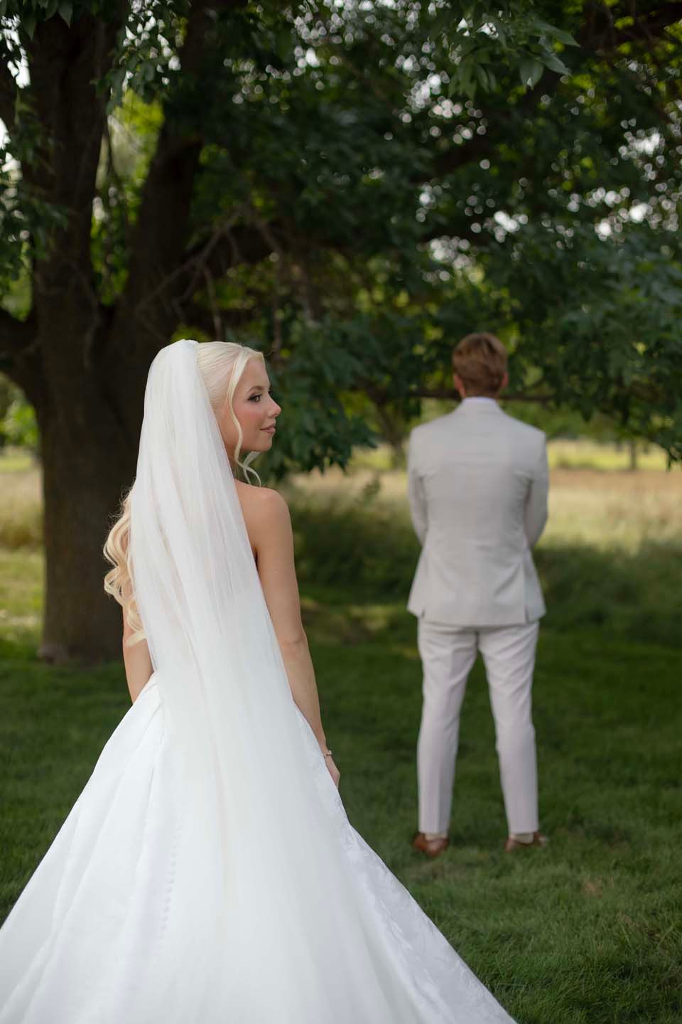 Bride in white gown and veil smiles as groom in light suit stands under green trees outdoors.