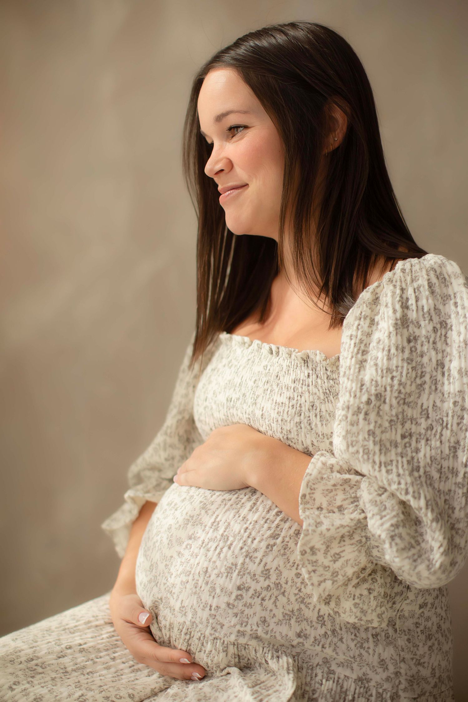 Smiling pregnant woman in floral dress cradling baby bump while sitting against neutral studio background.