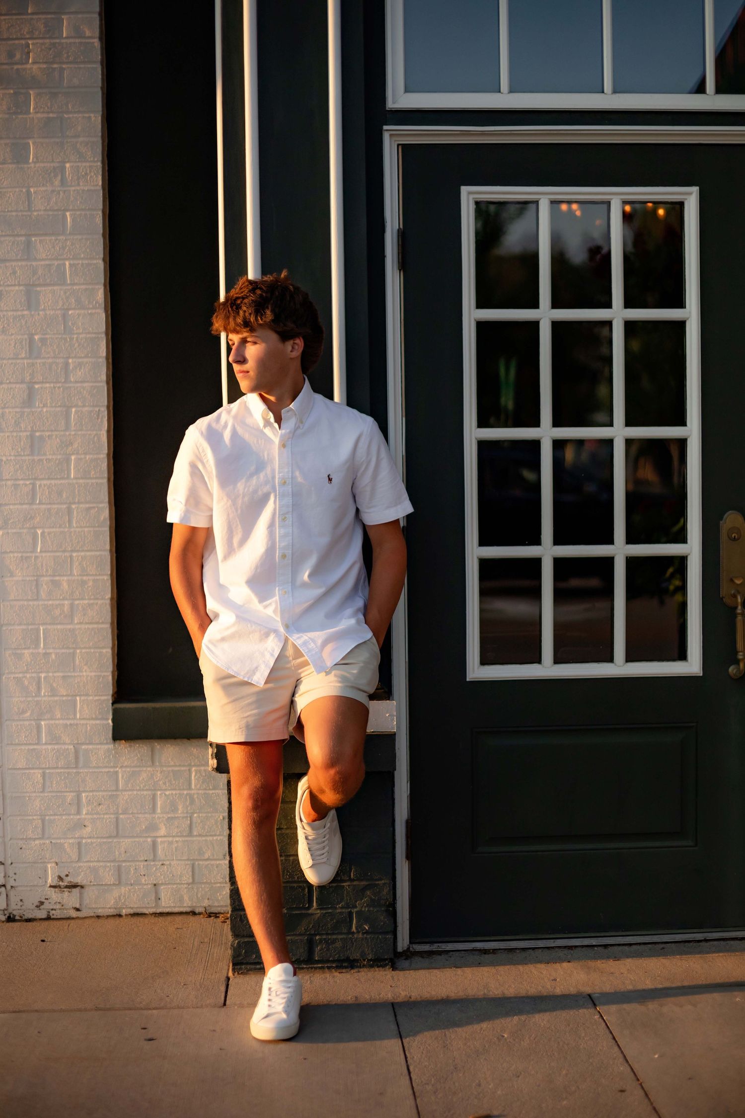Teen boy in white Polo Ralph Lauren shirt and shorts leaning against a dark green doorway at sunset.