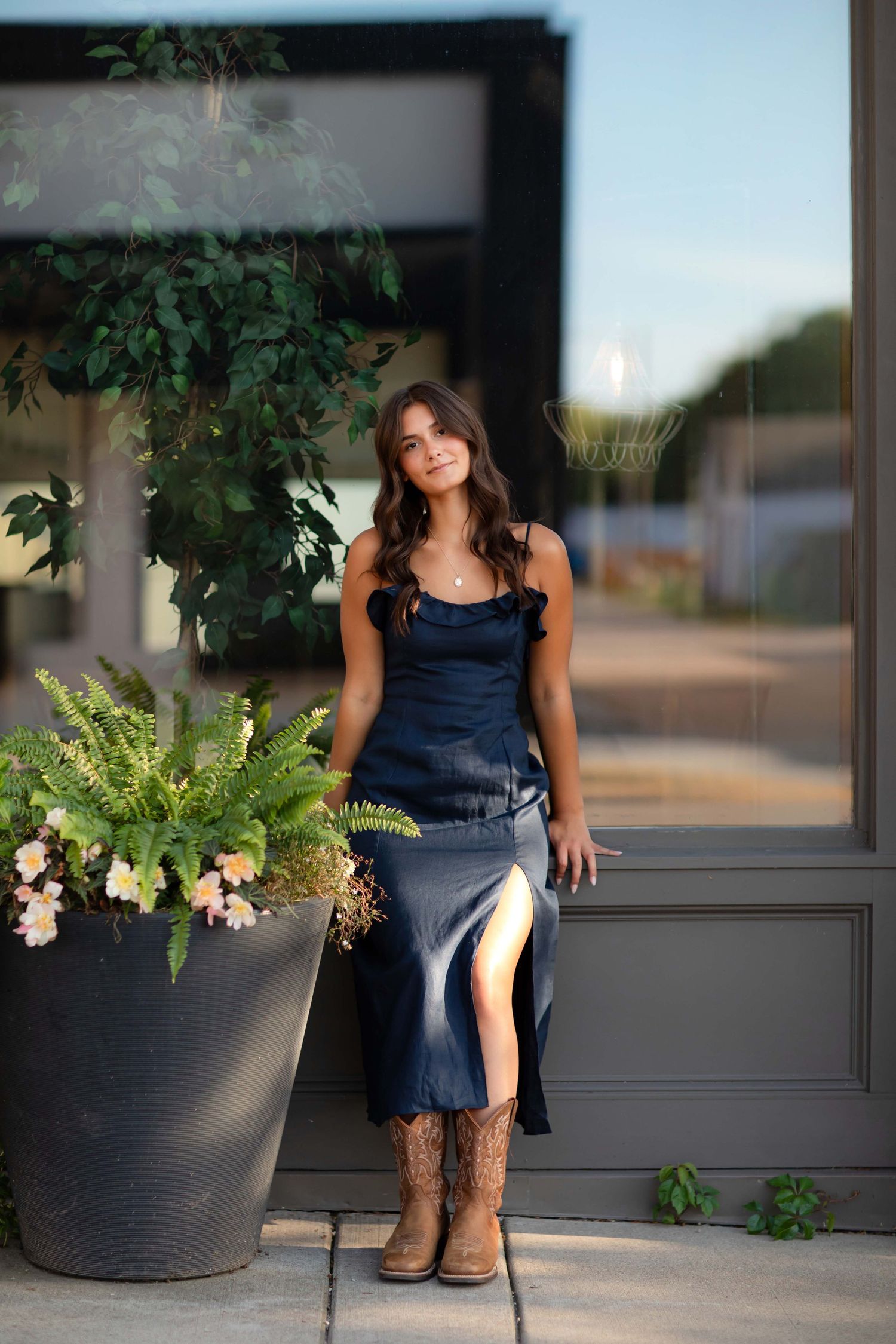 Young woman in navy slip dress and cowboy boots poses beside potted plants outside a modern building.