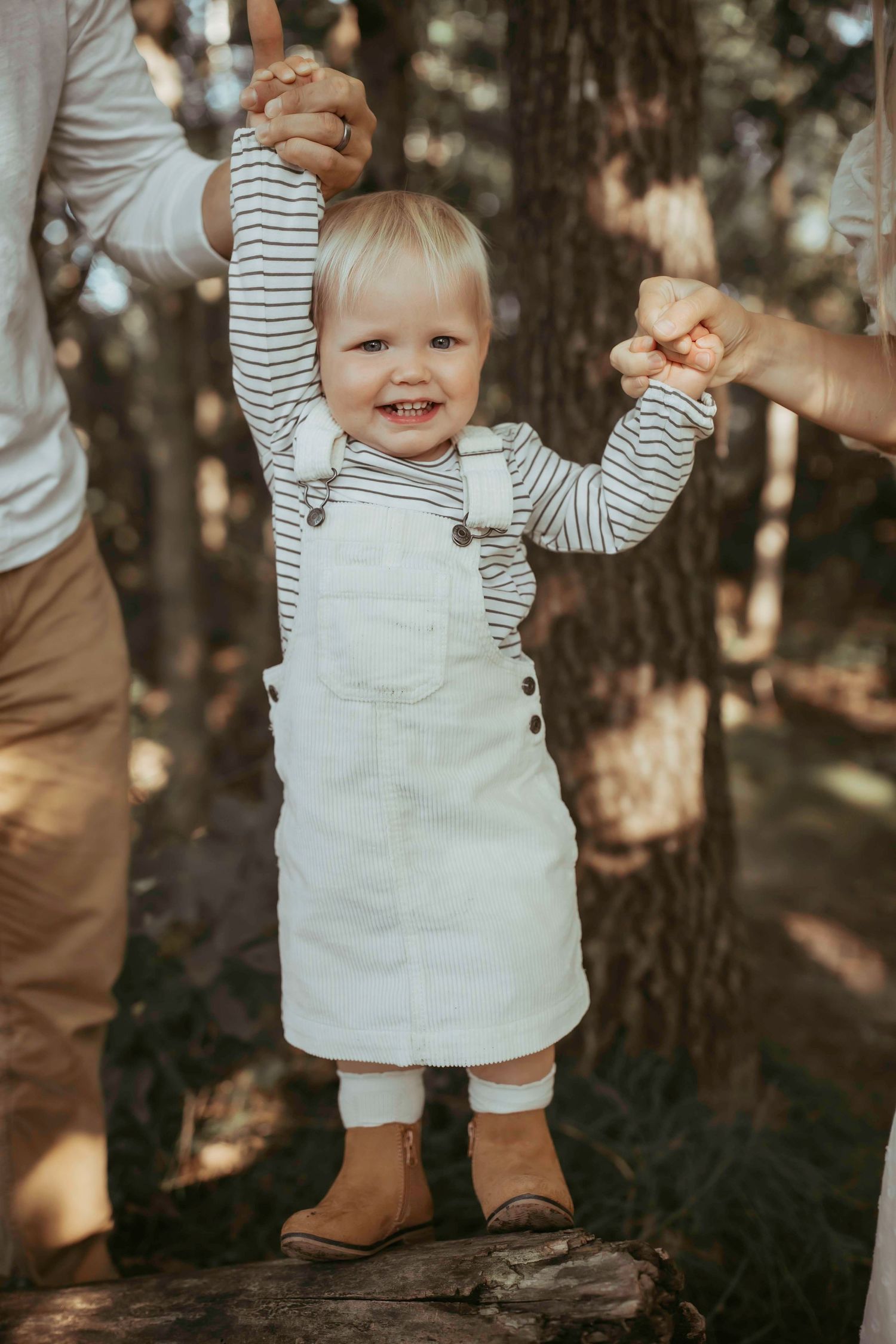 A toddler in white overalls and striped shirt smiles while being held by adult hands in a wooded setting.