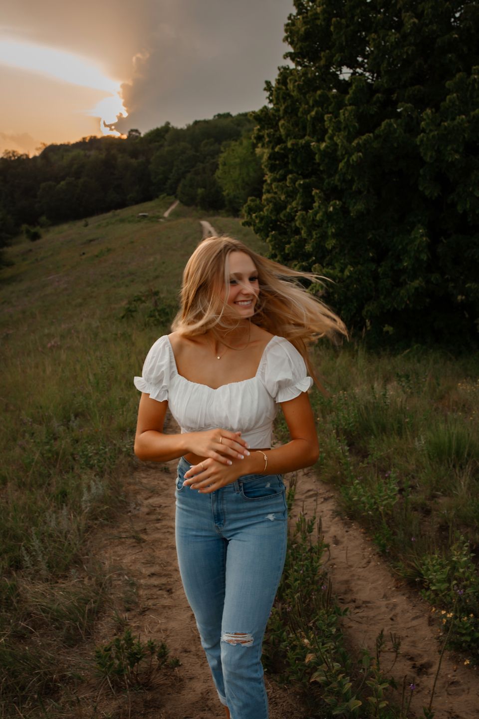 A woman wearing a white peasant top and jeans poses on a dirt path at sunset with windblown hair.