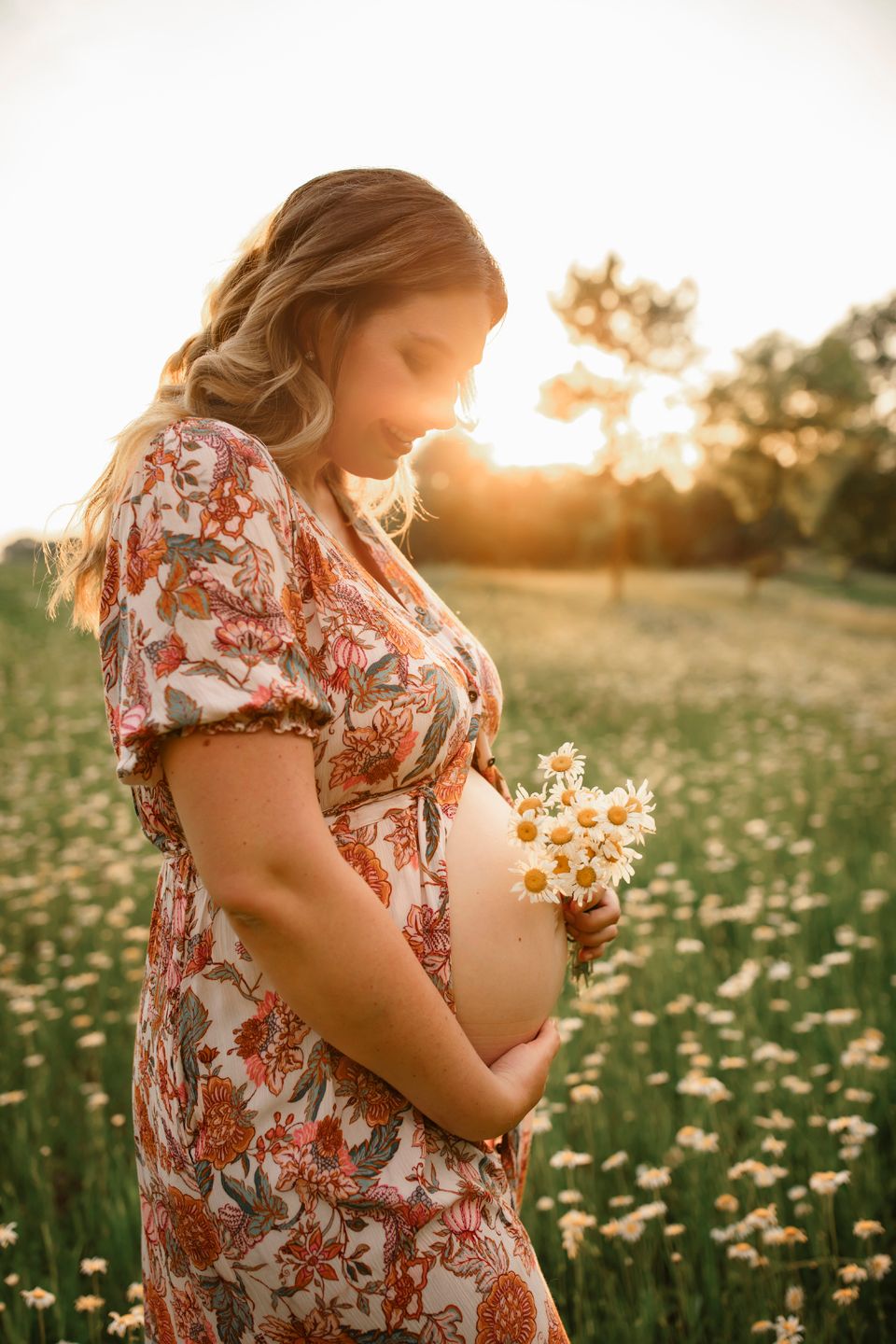 A sunset maternity photo shows a person in a floral dress holding wildflowers in a field of daisies.