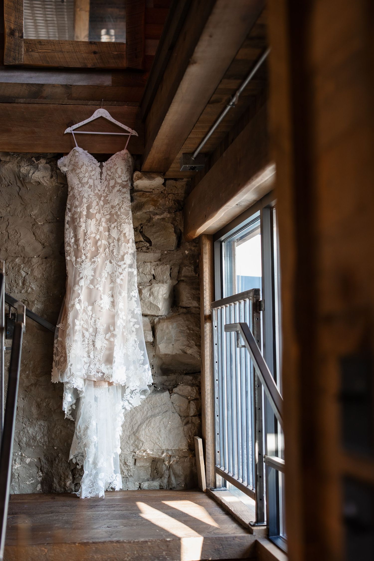 A delicate white lace wedding dress hangs against a rustic stone wall near a window.
