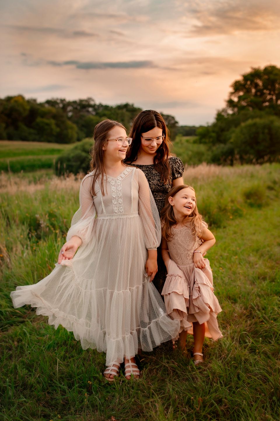 Three figures in flowing dresses share joyful moment in summer field.
