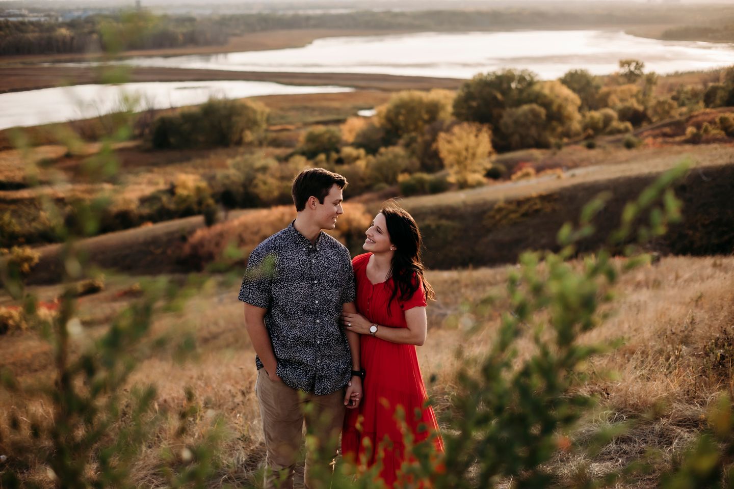 Two people share an intimate moment on a grassy hillside with a serene lake view at sunset.