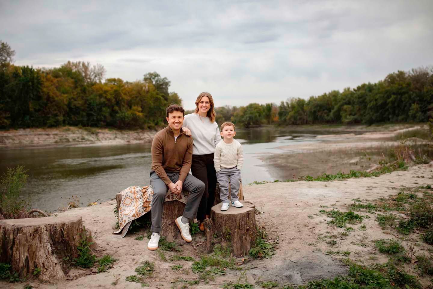 A family sits on tree stumps by a river on an overcast day.