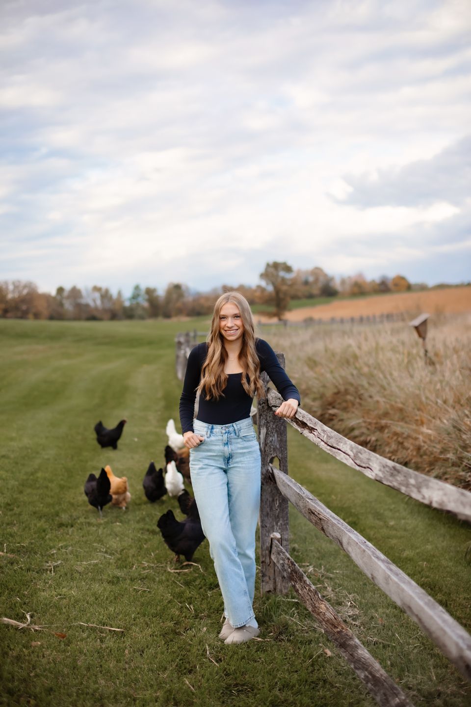 A casual autumn portrait in a rural setting with chickens grazing on green grass near a rustic wooden fence.