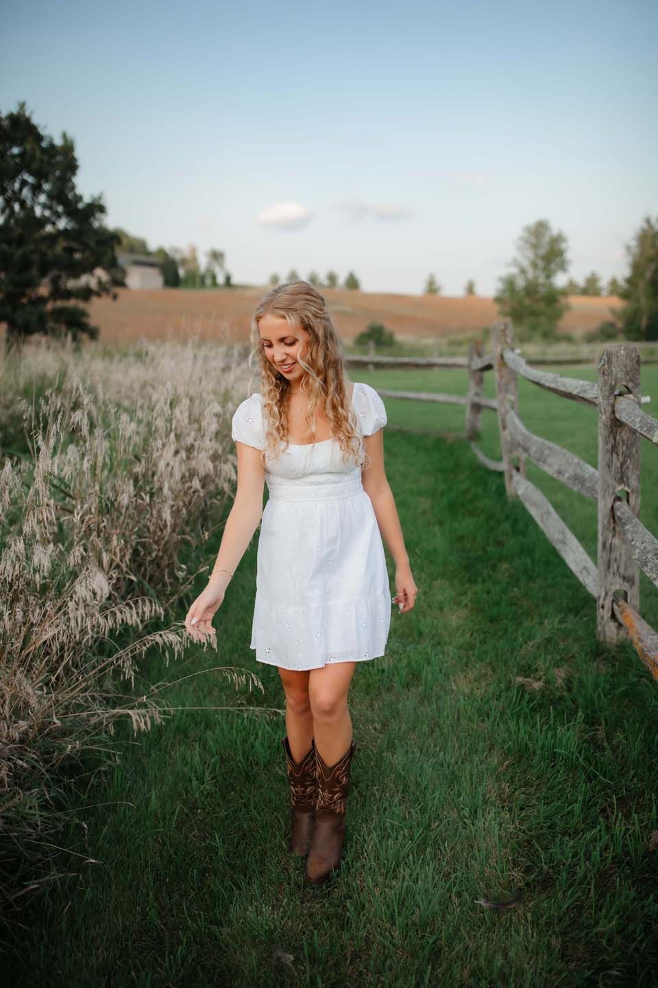 Casual countryside portrait with brown boots and white dress along wooden fence at sunset.