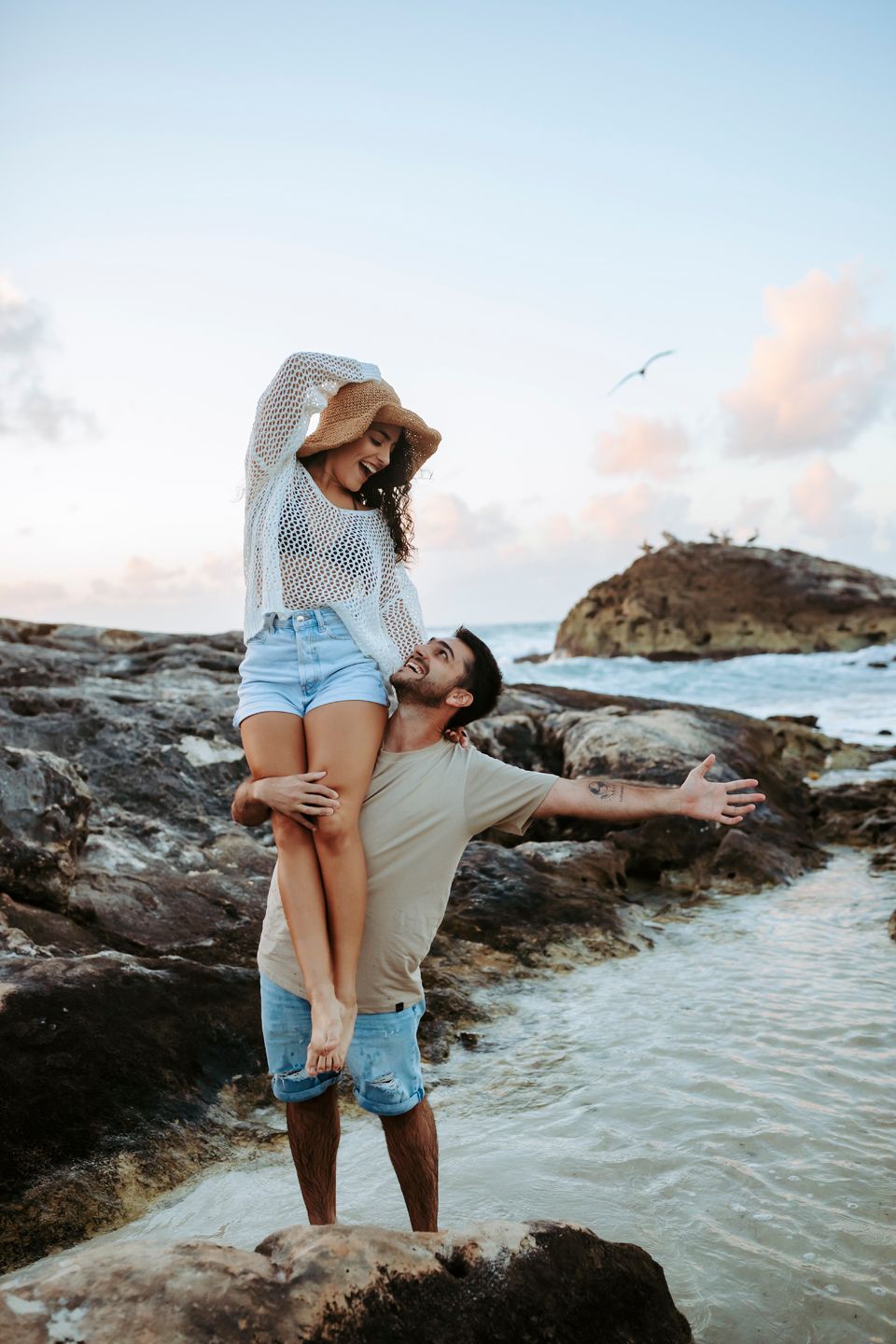 Playful couple posing on coastal rocks with ocean waves splashing behind them.