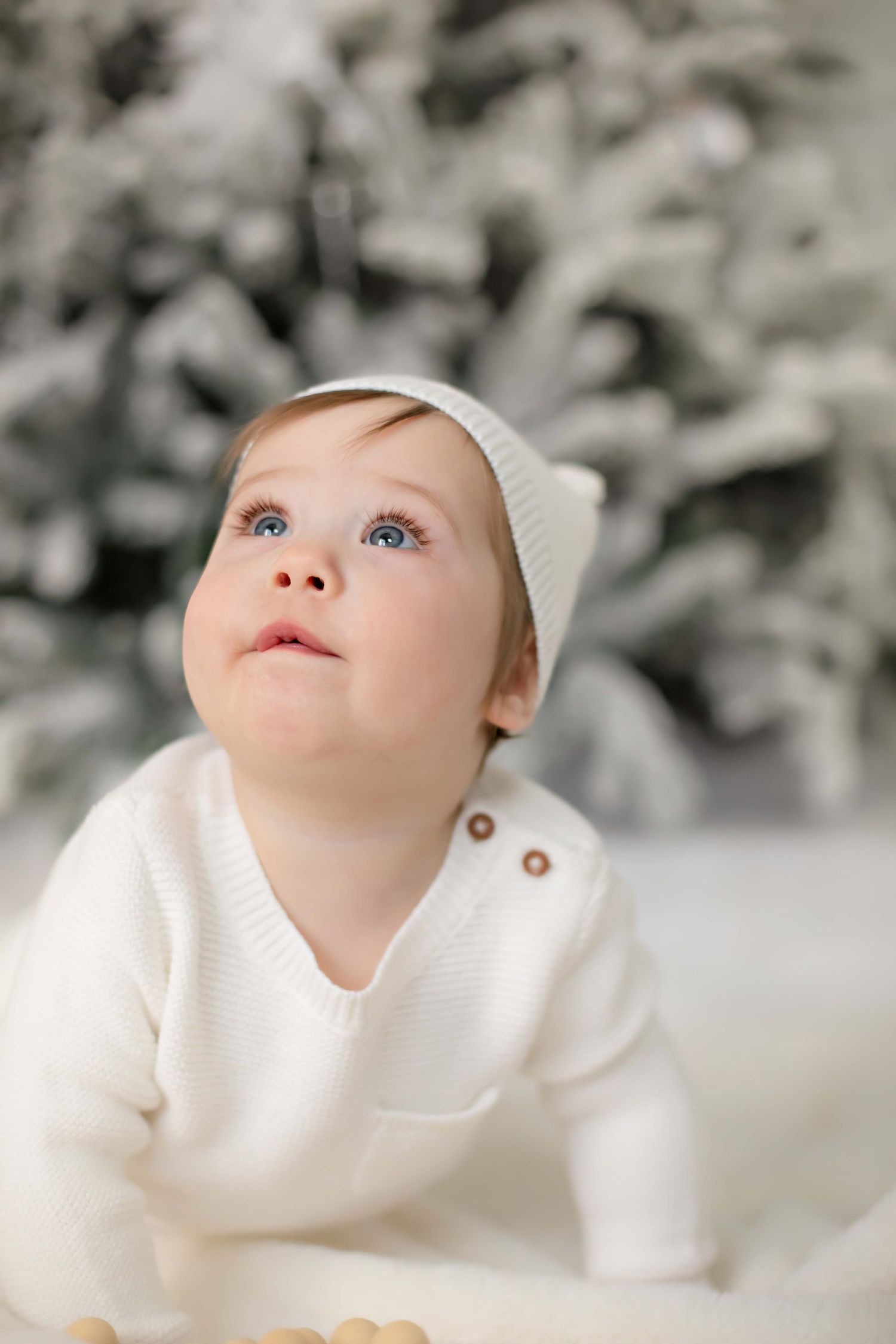 Small child in white sweater and beanie gazes up against snowy winter backdrop.