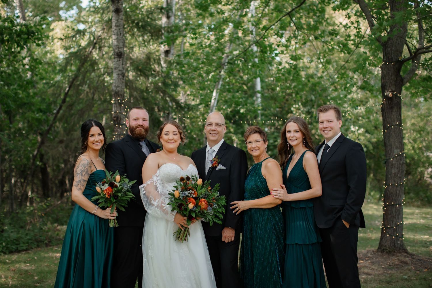 Wedding party in emerald green dresses posing for a formal group photo in a wooded setting.