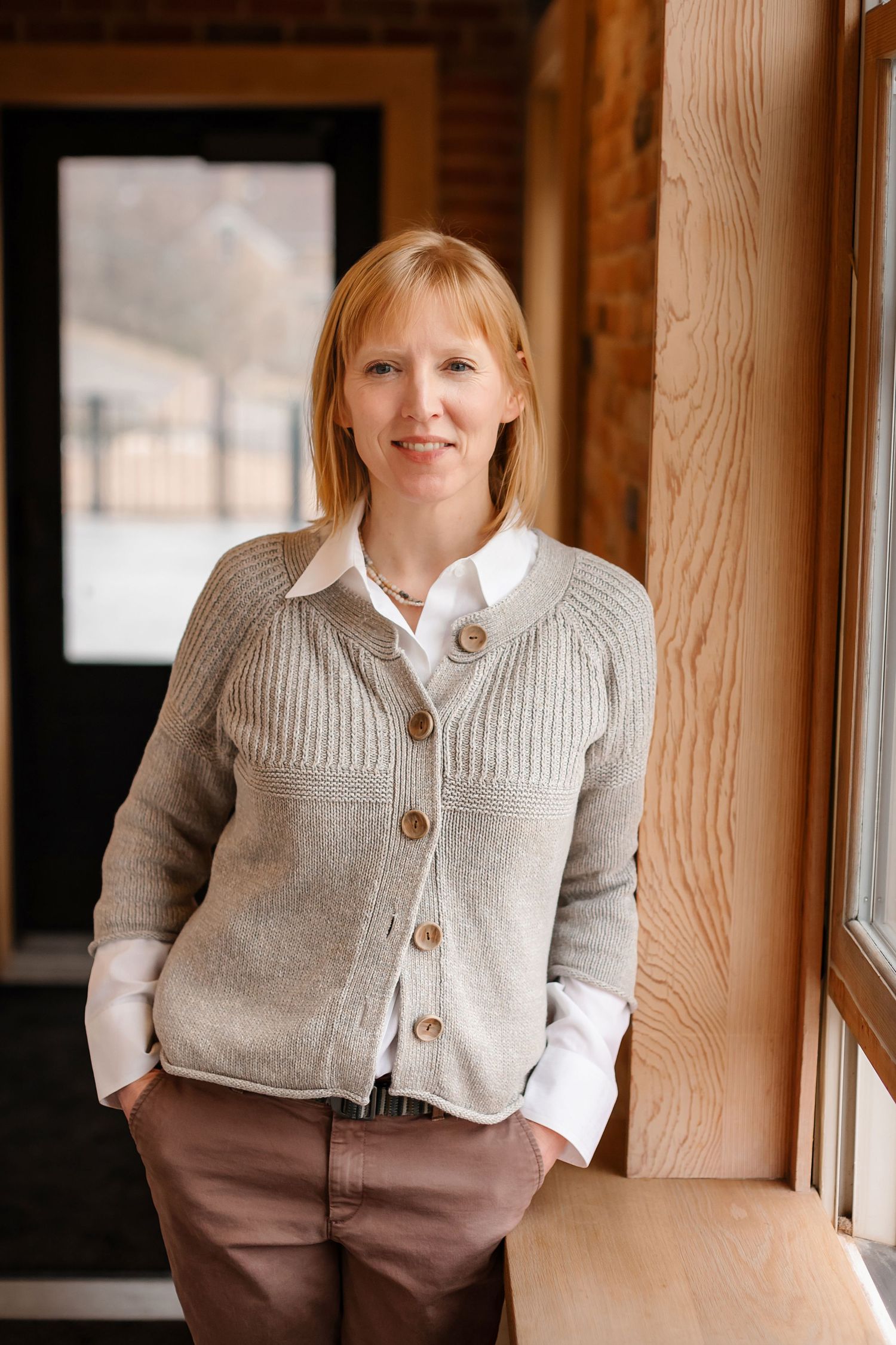 Professional portrait of person in cozy gray cardigan and khaki pants by wooden window frame.