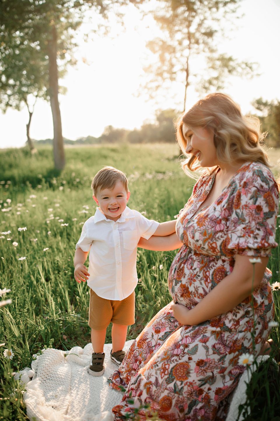 A floral dress-clad woman and young child share a joyful moment on a picnic blanket in a sunlit meadow.