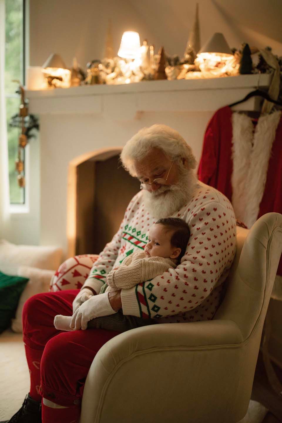 Santa Claus in festive pajamas holds a sleeping baby in an armchair by a decorated fireplace.