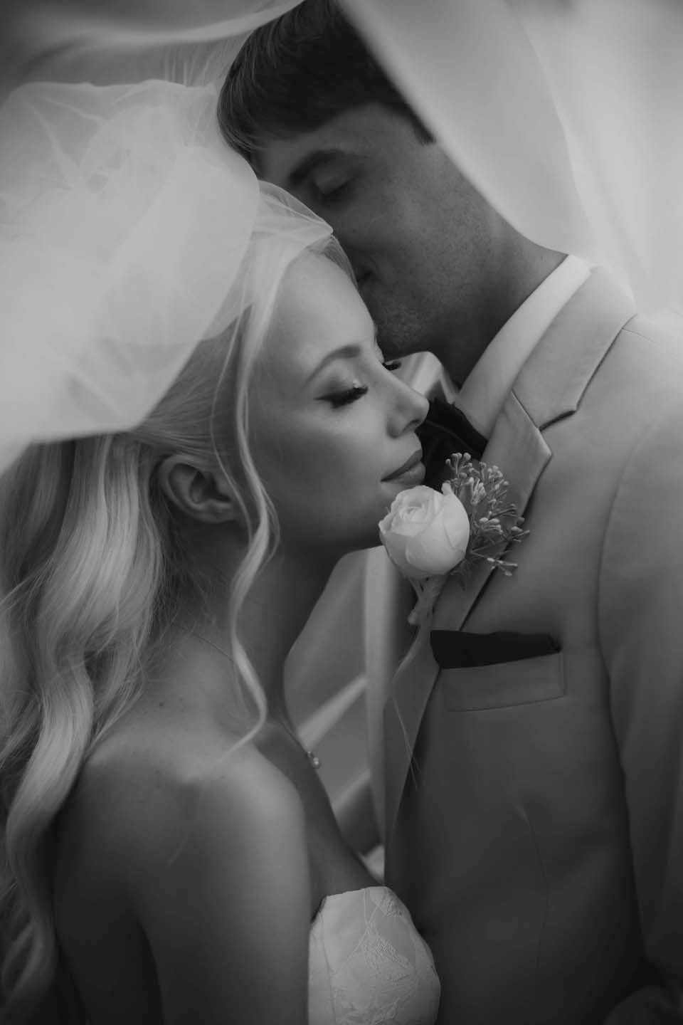 Black and white wedding photo of bride and groom sharing intimate moment under veil, groom kissing bride's forehead.