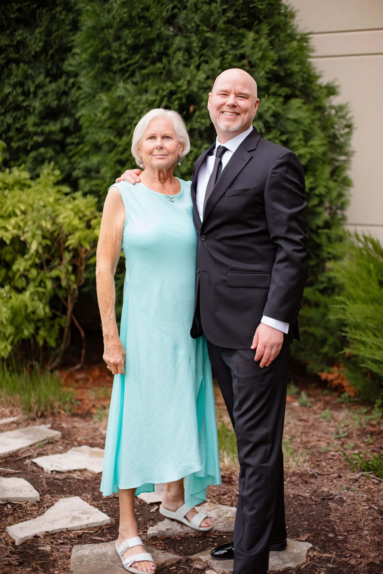 Couple in formal attire posing together in a garden setting with green shrubs in the background.