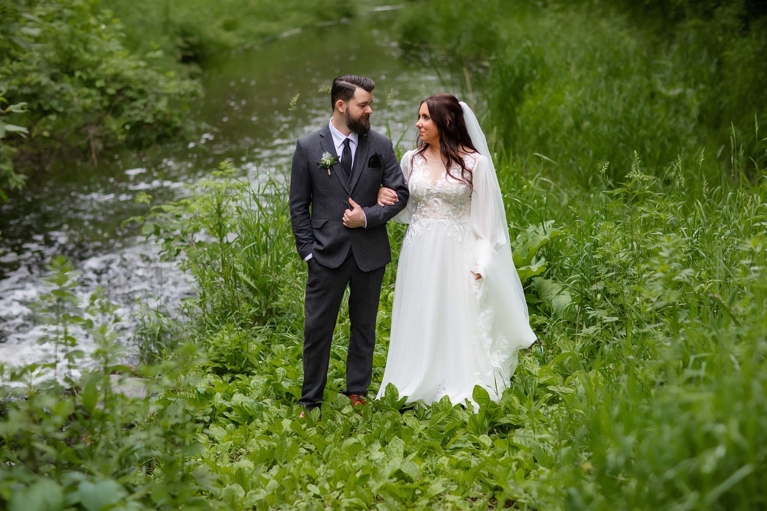 A bride and groom, intimate moment while standing in lush green grass near a flowing stream. Alexandria, Mn with Willow & Wren Photography.