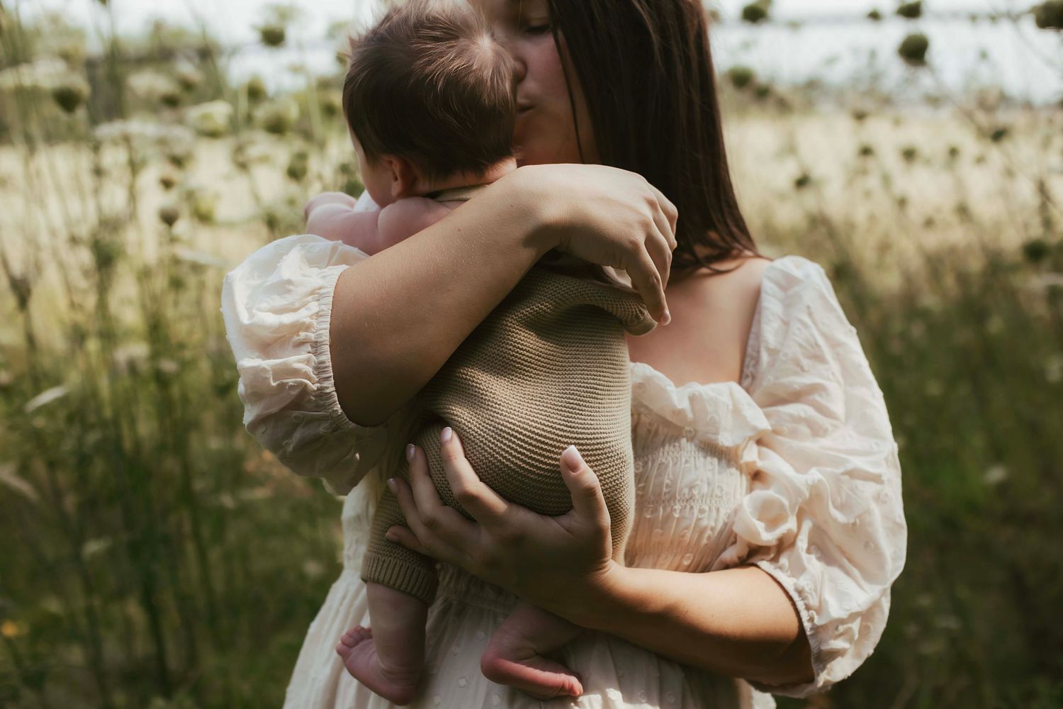 A mother in a white dress tenderly holds her newborn baby in a sunlit wildflower field.