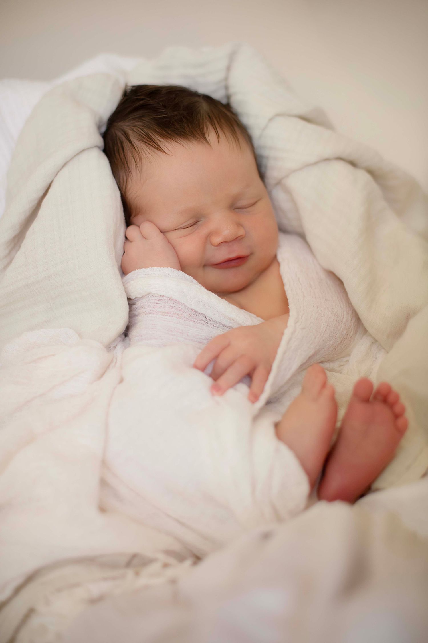Peaceful newborn baby sleeping wrapped in soft white blanket with tiny feet visible and gentle smile.