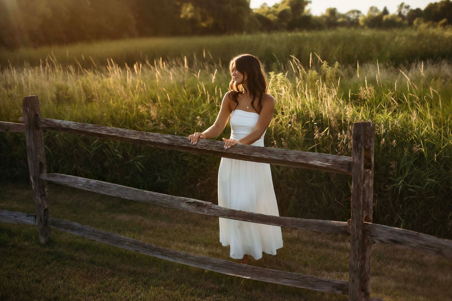 Woman in white dress leaning on rustic wooden fence in golden hour sunlight with lush green field backdrop.