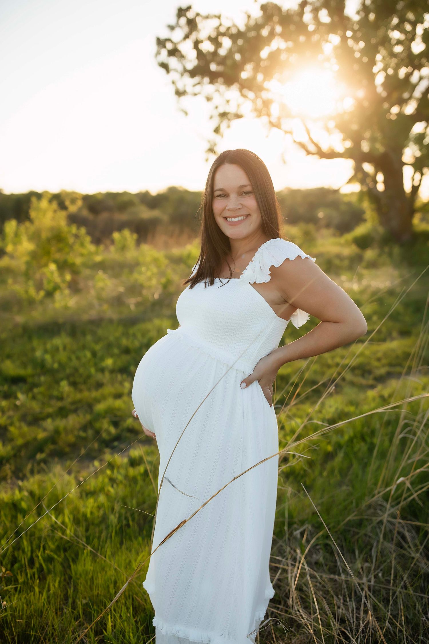 Smiling pregnant woman in white dress holding baby bump outdoors at golden sunset in green field.