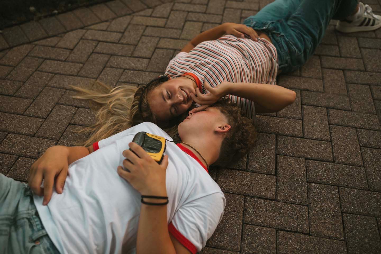 Two young friends lying on brick pavement, smiling and holding a yellow device, enjoying a carefree moment together.