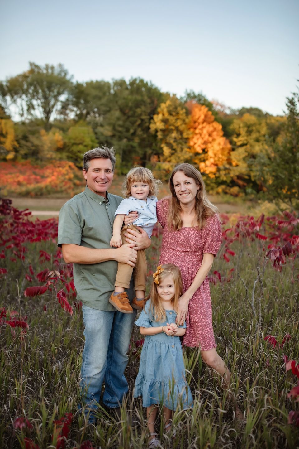 Family poses together in autumn field with vibrant red foliage and golden trees in background at sunset.
