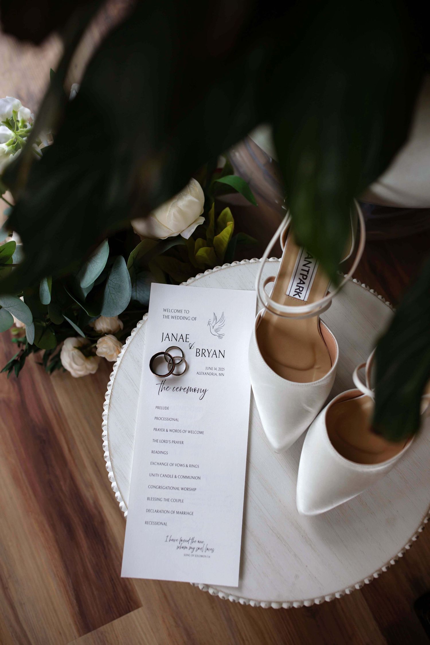 Wedding details including white bridal shoes and ceremony program with rings design on a round table.