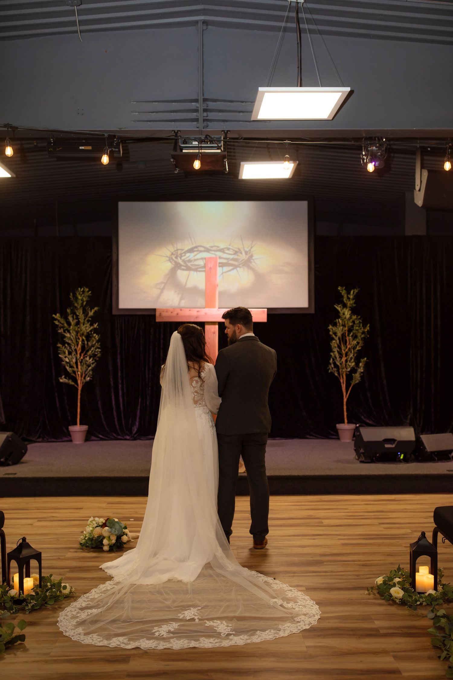 A long white wedding dress train flows across a wooden floor in a dimly lit venue with lanterns and decorative trees.