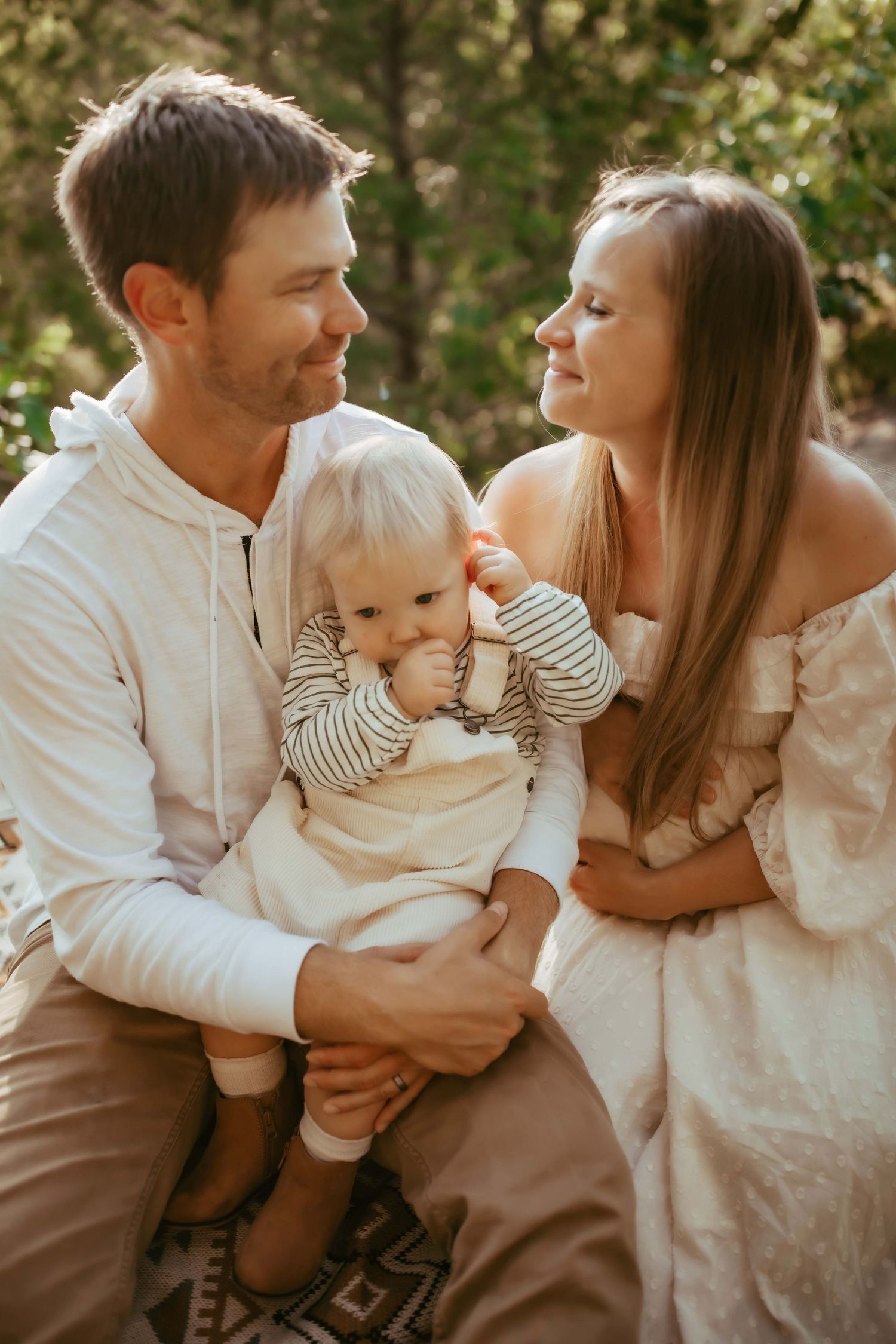 An intimate family portrait shows a couple in white clothing sharing tender moments with their baby on a patterned blanket.