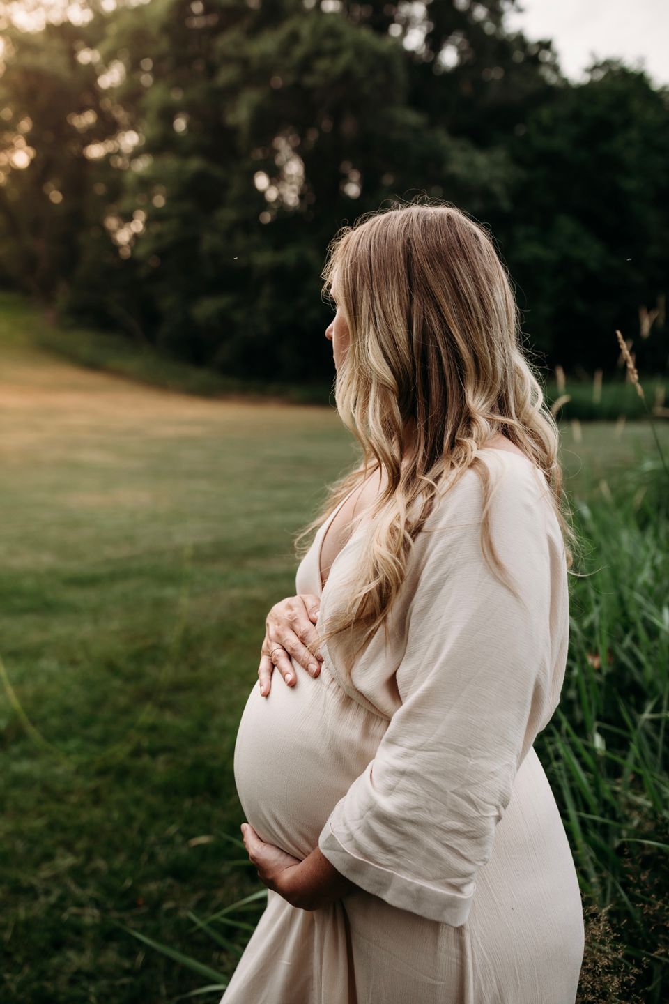 A pregnant woman in a cream dress cradles her baby bump during a sunset photoshoot in a grassy field.