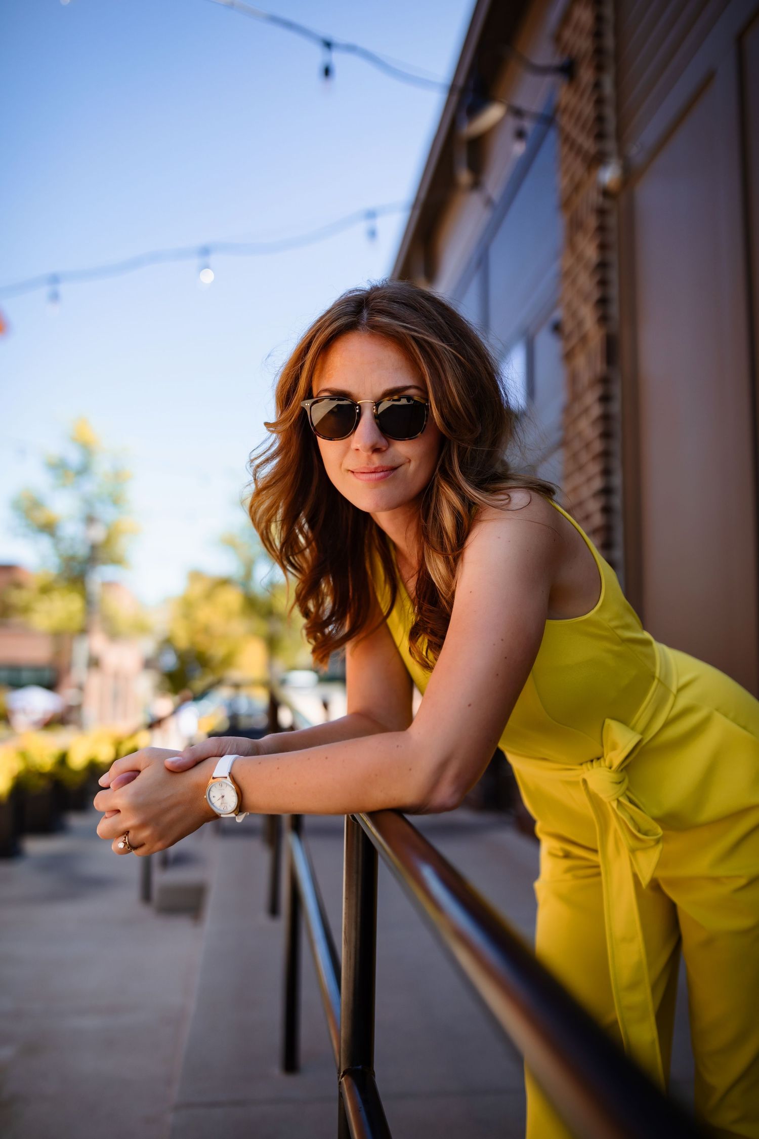 Woman in bright yellow dress leans on railing at sunset with string lights in background.
